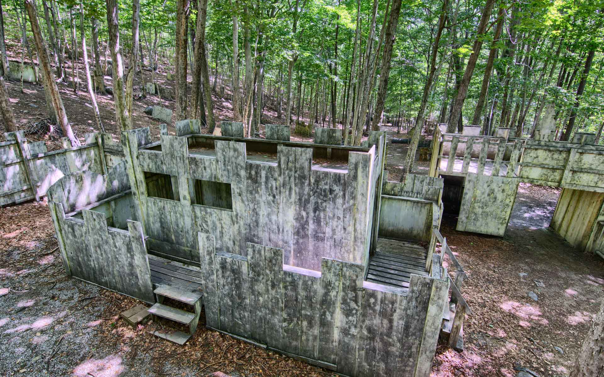 Paintball course with wooden structures in a forest. Grey structures, green trees.