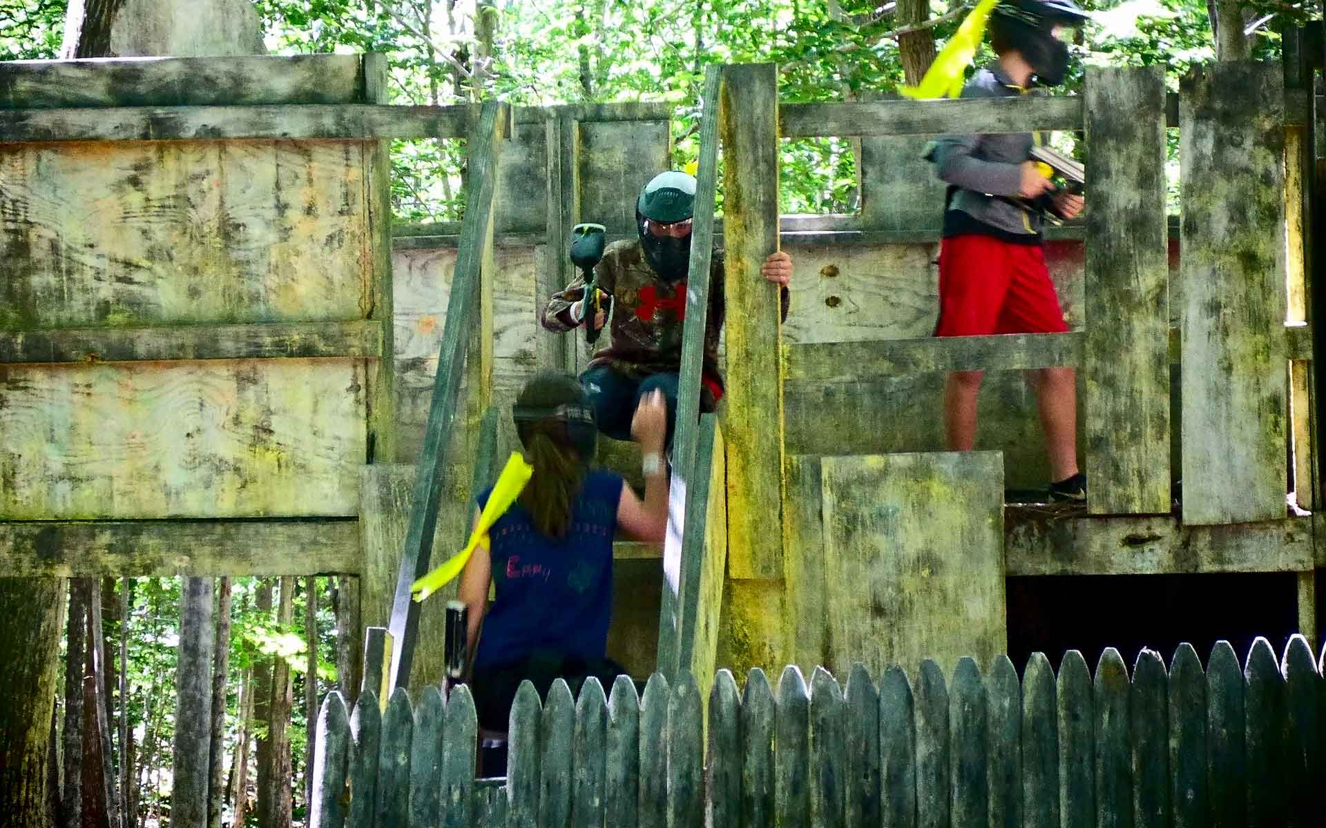 People playing paintball behind wooden barricades in a forest, some holding guns.