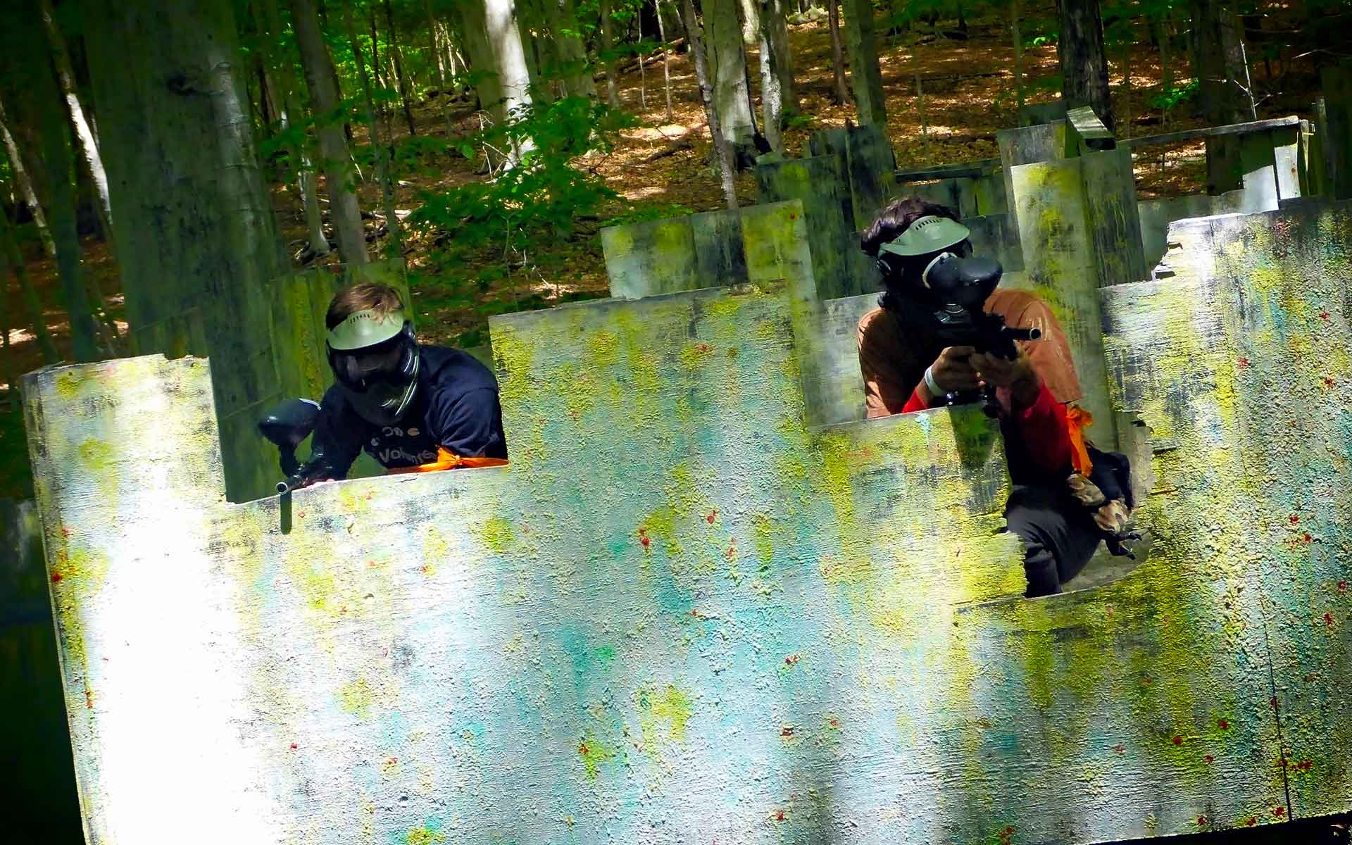 Two people in paintball gear aiming guns from behind a concrete wall in a wooded area.