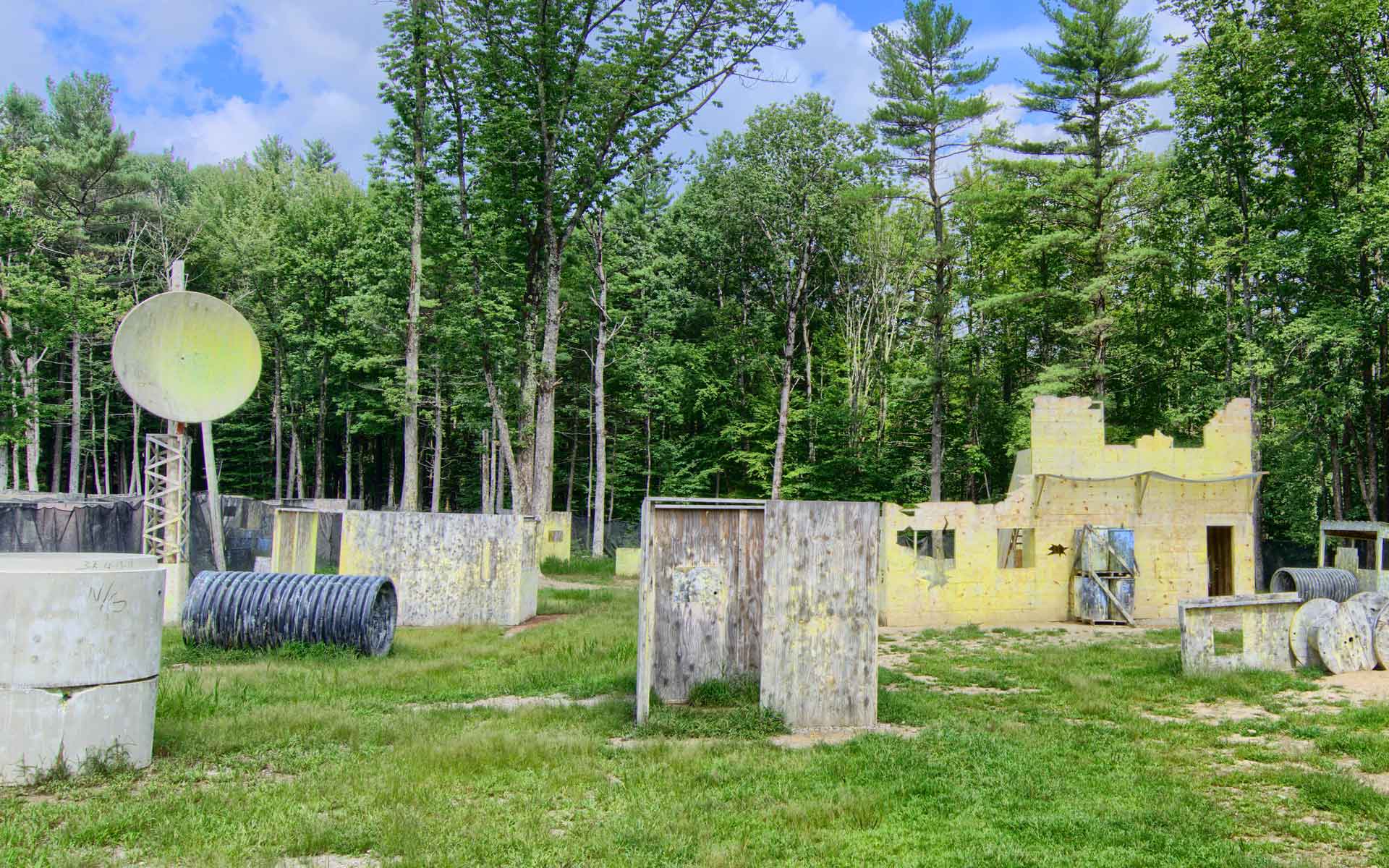Paintball field with bunkers and a yellow building facade against a backdrop of trees.