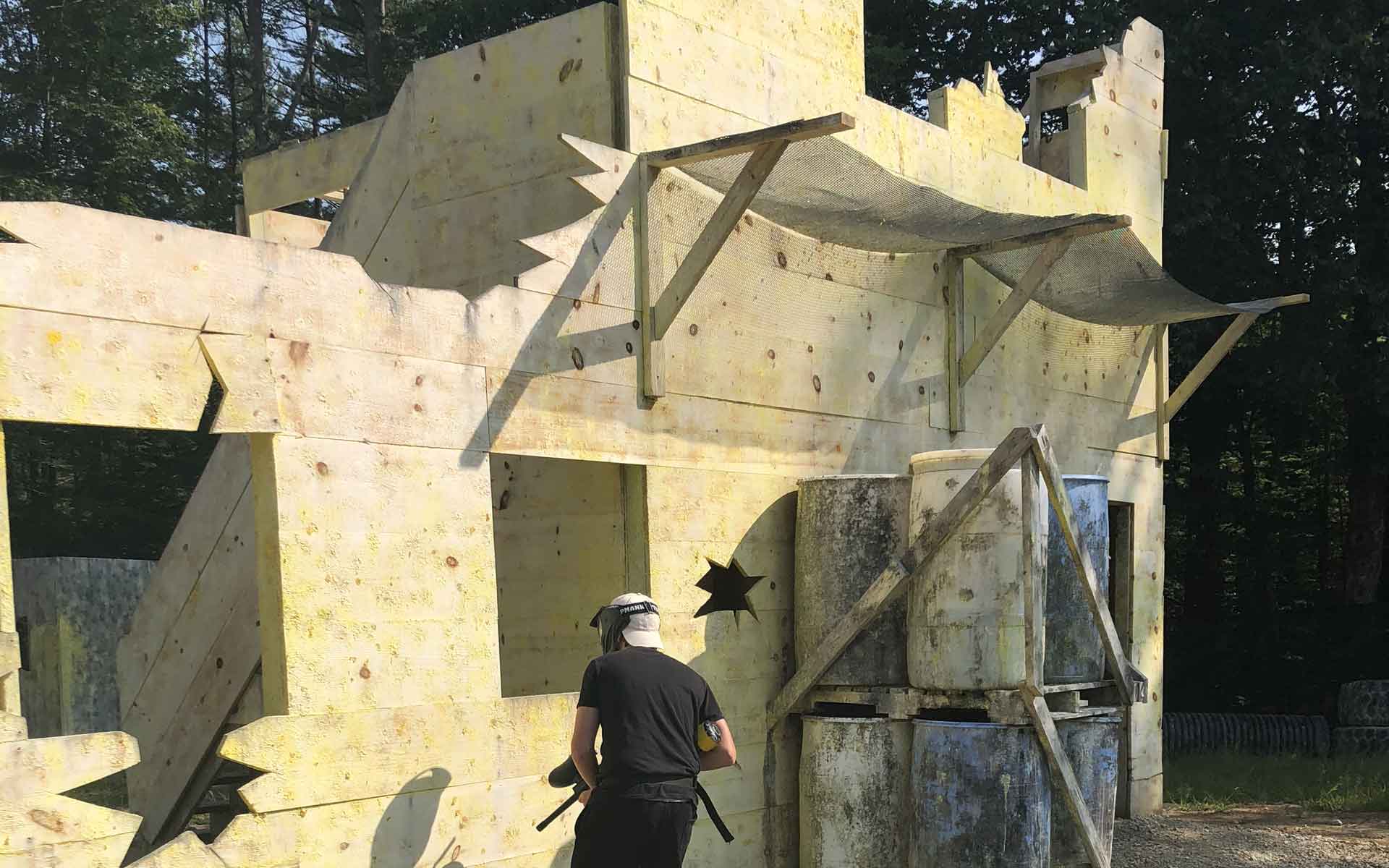 Person playing paintball in a partially destroyed, yellow building; surrounded by trees.