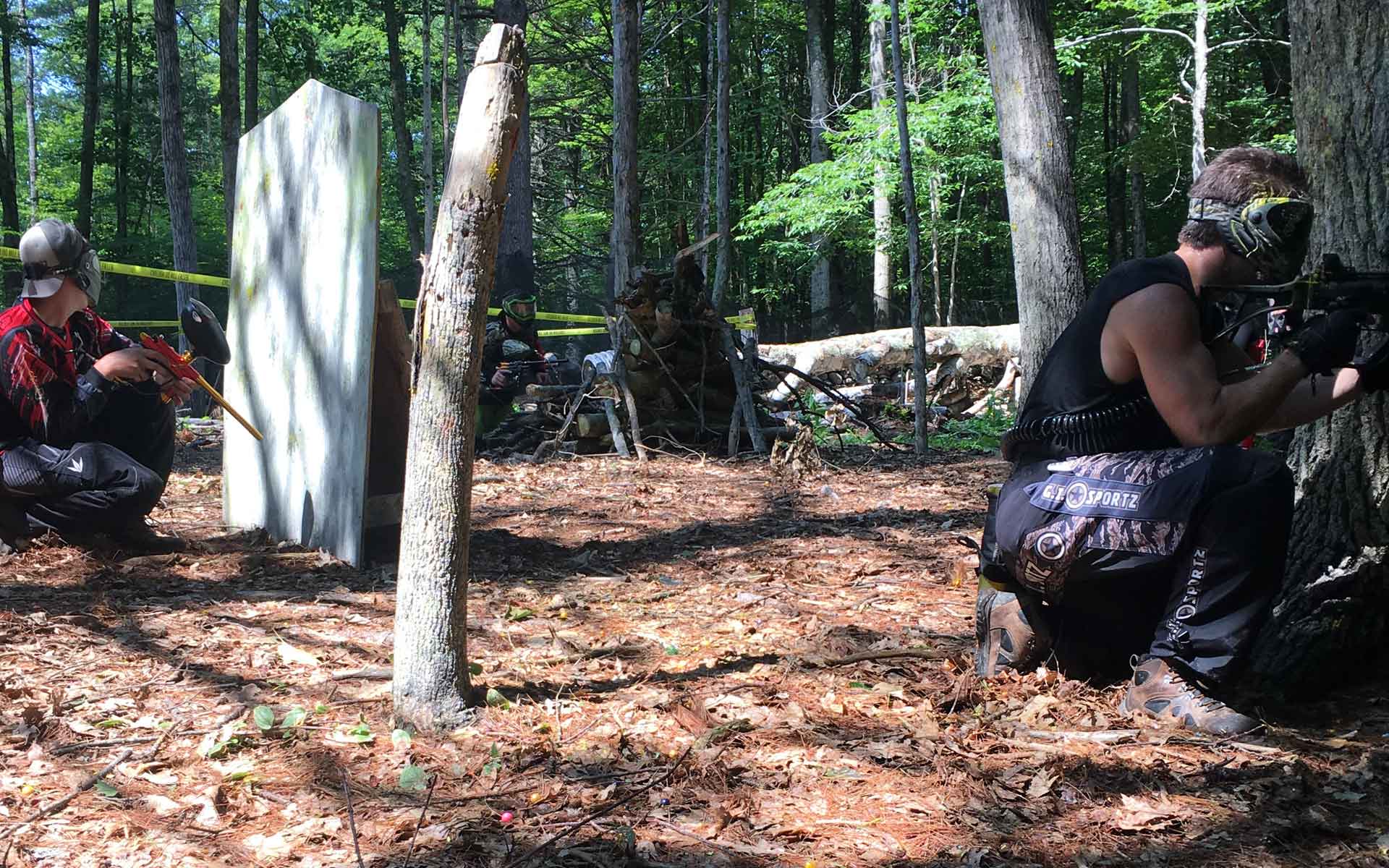 Two people in paintball gear crouch in a wooded area, aiming paintball guns.