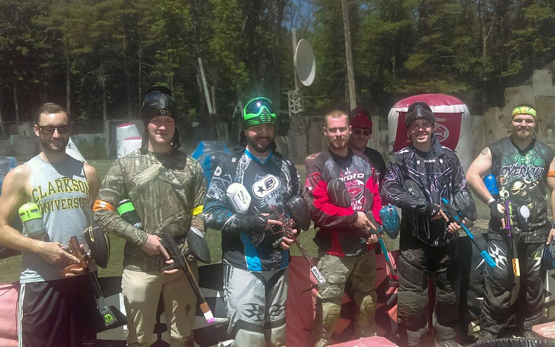 Group of paintball players pose outdoors, wearing protective gear and holding markers. Sunny day.