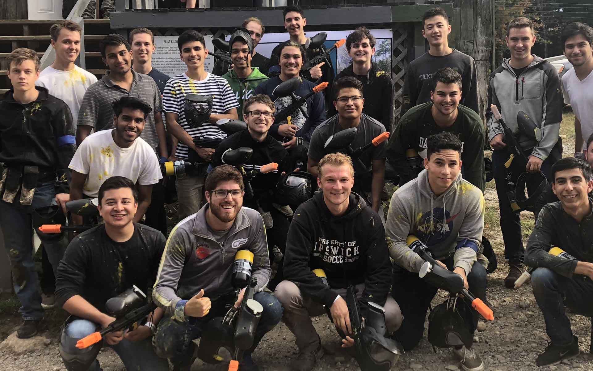 Group of young men in paintball gear, posing outdoors. They are holding paintball guns and smiling.
