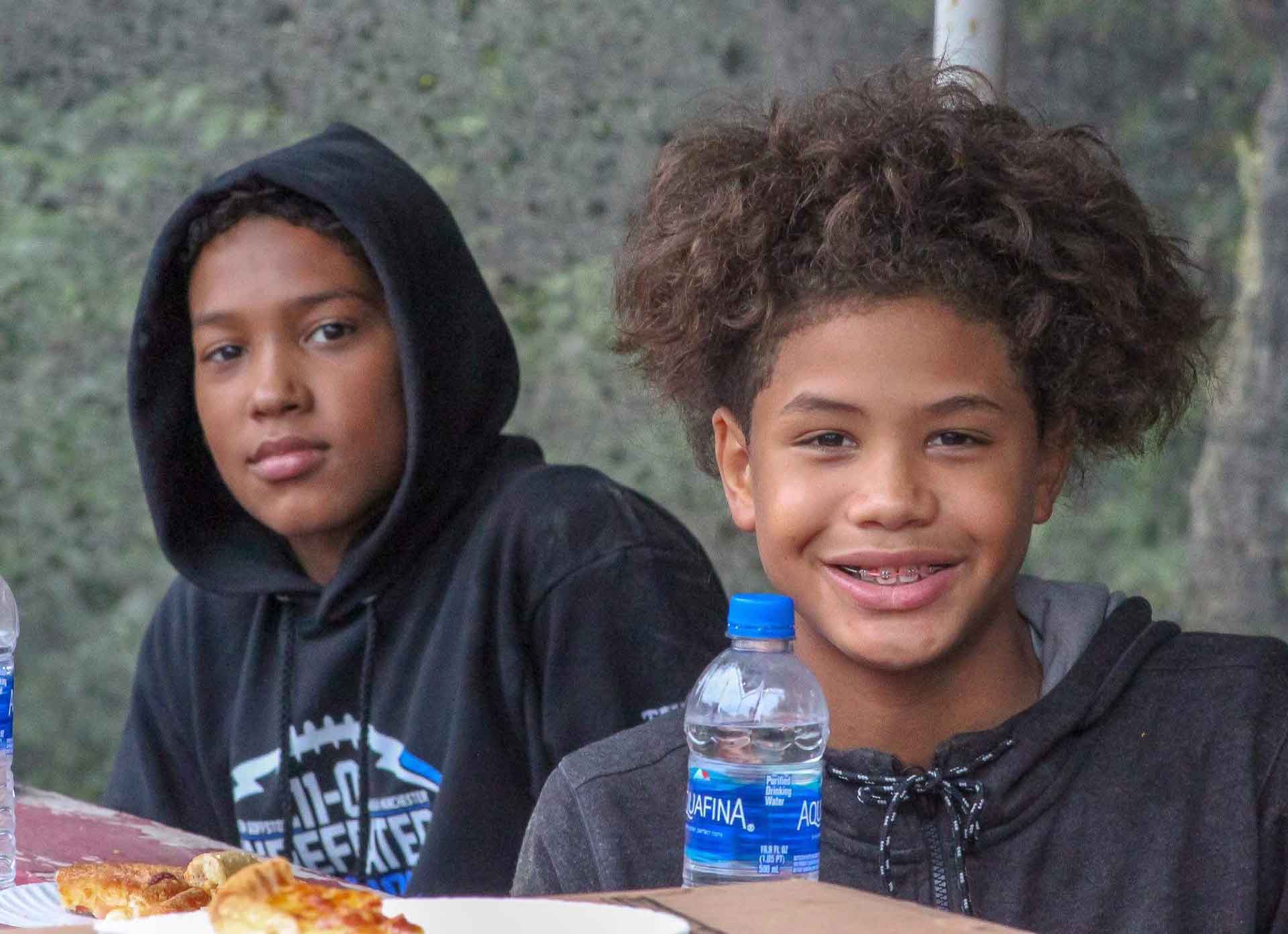 Two young boys smiling at a picnic table. One has curly hair and braces. Both are wearing hoodies.