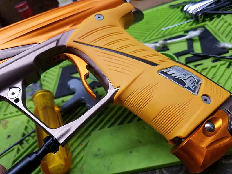 Close-up of an orange and silver paintball gun being worked on; detailed view of grip and trigger.