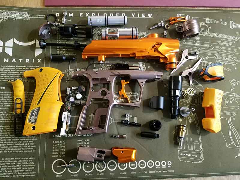 Disassembled paintball gun parts in orange and purple on a green mat.