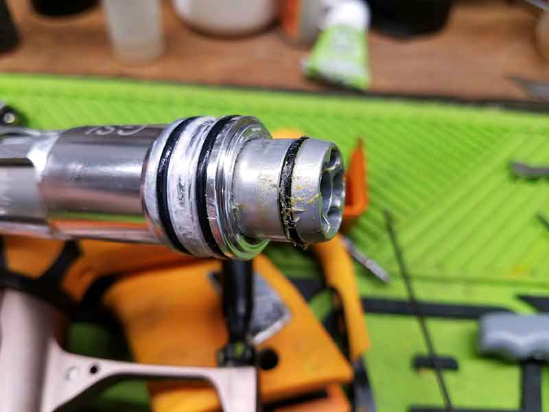 Close-up of a silver paintball marker part with black and white seals, showing wear and tear with yellow residue.