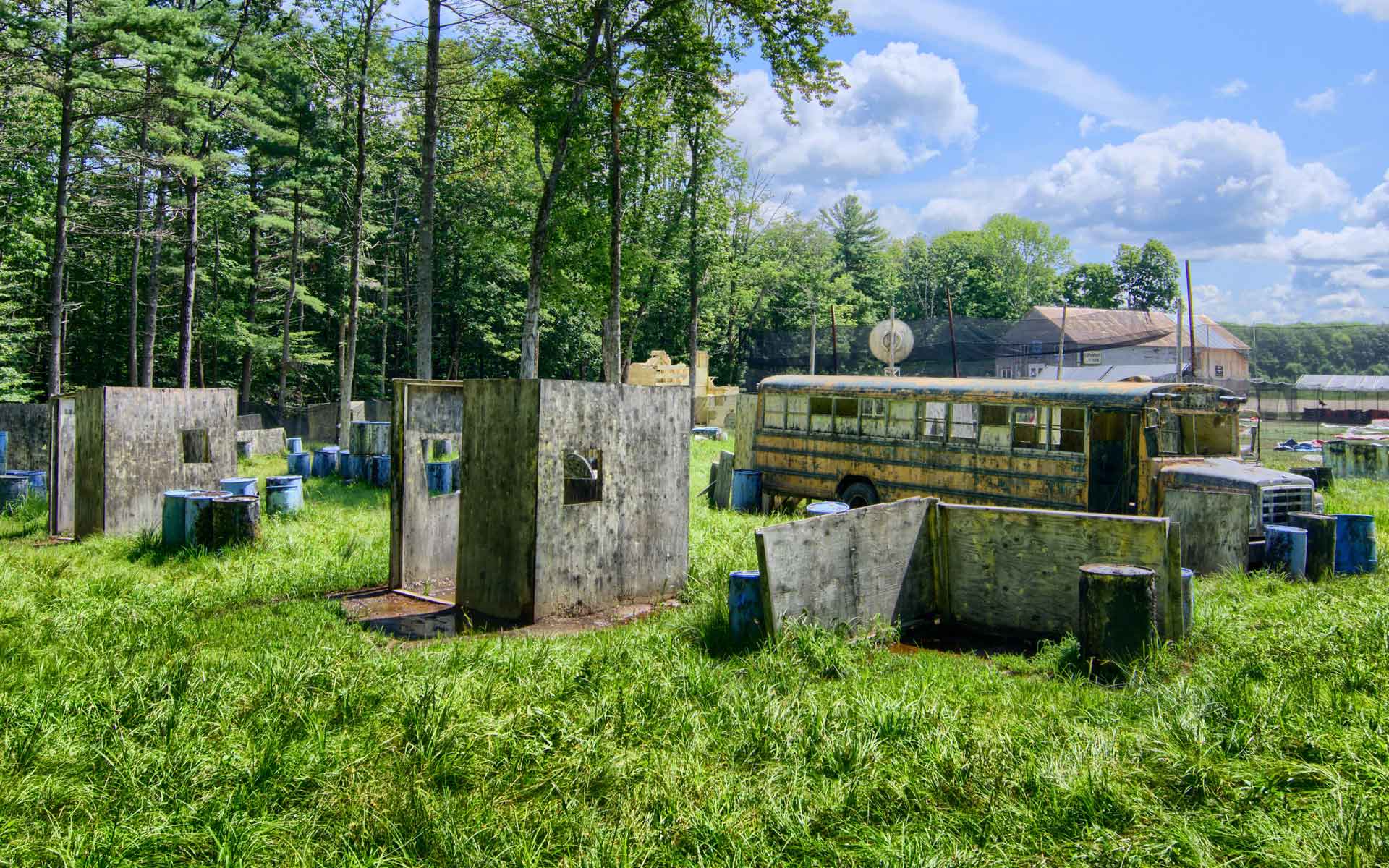 Paintball field with wooden bunkers, blue barrels, and an old yellow school bus in a grassy area.