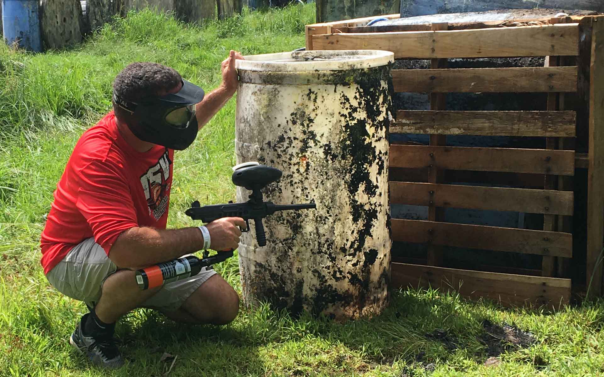 A person kneels, aiming a paintball gun behind a barrel and wooden structure.
