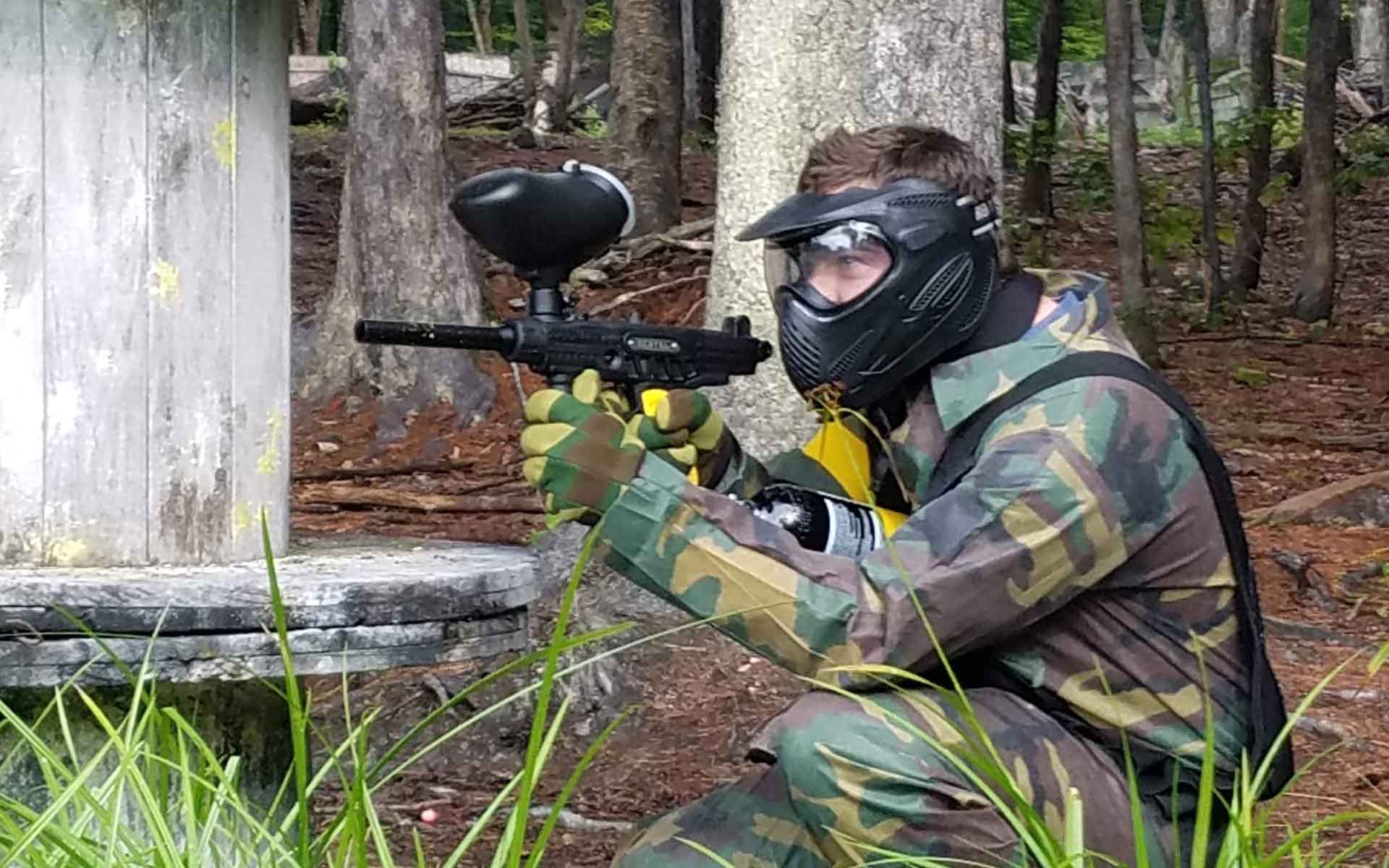 Person in camouflage gear aiming a paintball gun in a wooded area, crouching near a wooden bunker.