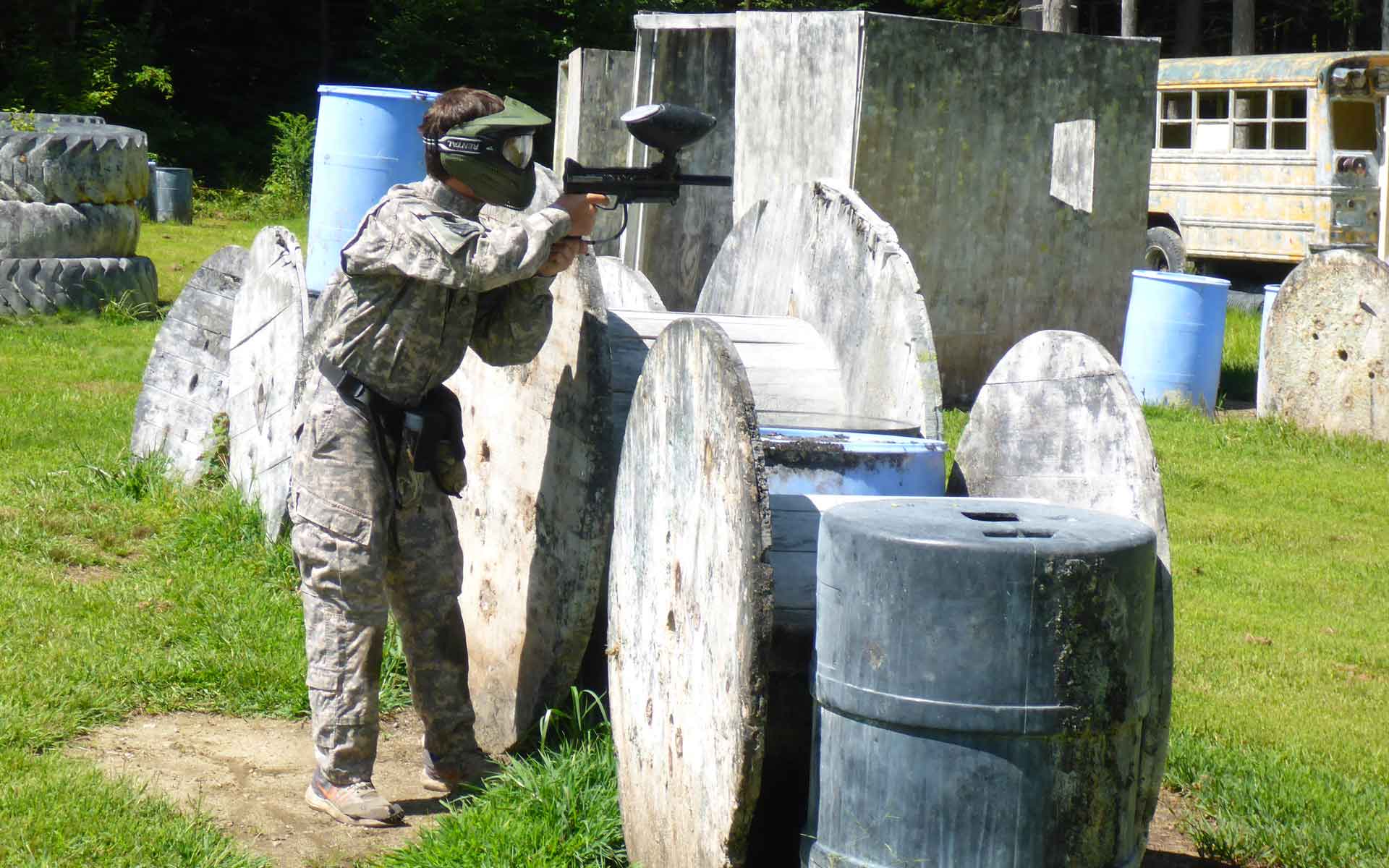 Person in camouflage aiming a paintball gun behind a barricade at a paintball course.
