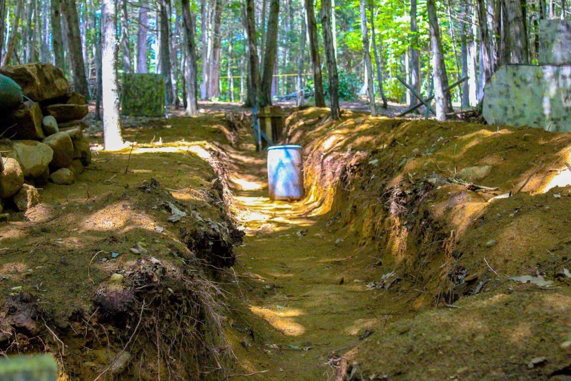 A dirt trench with a blue and white barrel in the middle, set in a wooded area.