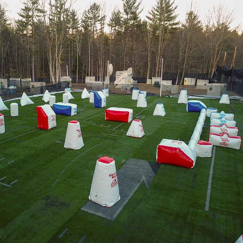 Paintball field with inflatable bunkers on artificial turf, surrounded by trees.