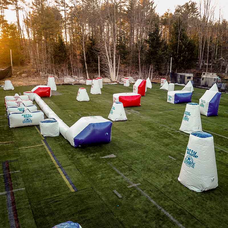 Paintball field with inflatable bunkers; red, blue, and white; set on artificial turf in a wooded area.