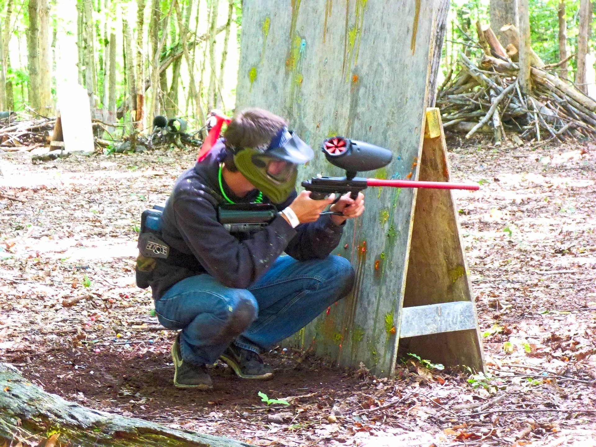 A person in paintball gear aims a gun behind a wooden barrier in a wooded area.