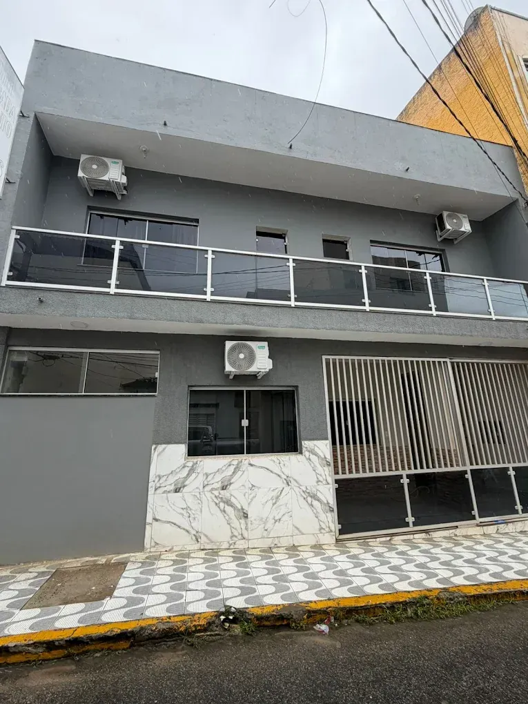 Prédio cinza com varanda de vidro, ar-condicionado e entrada com piso de azulejo, localizado em uma rua.