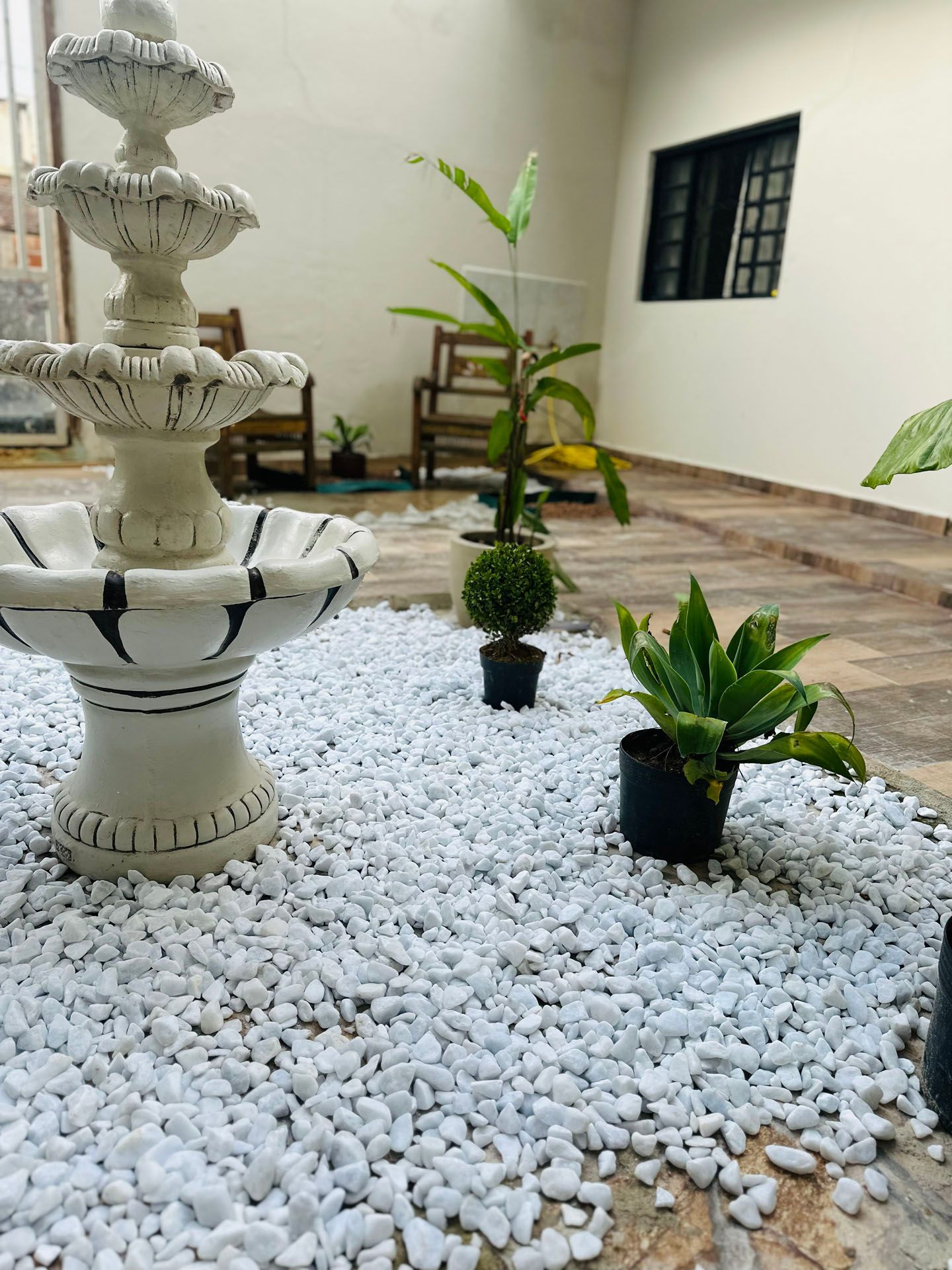 Pátio com fonte decorativa, cascalho branco e vasos de plantas.