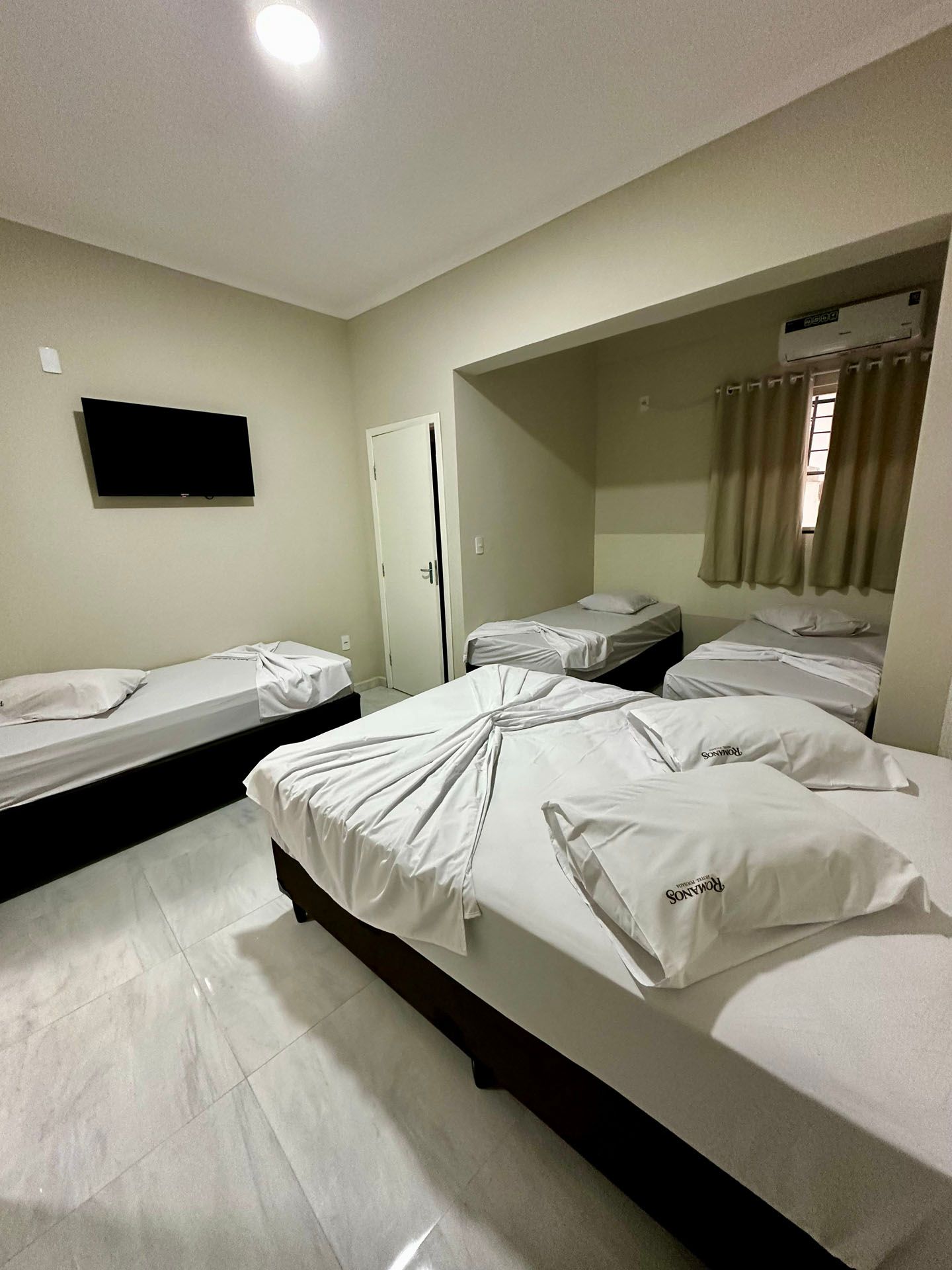 Um quarto de hotel com cinco camas, uma TV e uma porta fechada. Paredes e roupa de cama em tons de cinza claro.