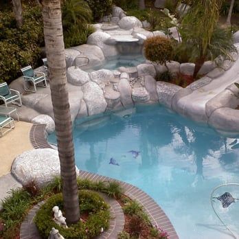 Swimming pool with a slide, waterfall, and spa. Palm tree in foreground, chairs on the deck.