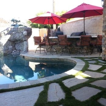 Poolside patio with bar, red umbrellas, stone features, and green grass accents.