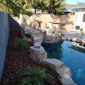 Rock waterfall feature beside a swimming pool, with landscaping. Sunny outdoor setting.