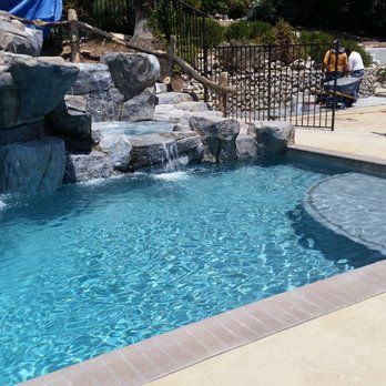 Pool with waterfall, spa, and rock features. Turquoise water, tan deck, black fence in background.