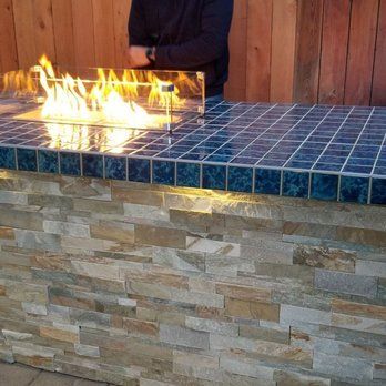 Outdoor fire pit built into a tiled counter and stacked stone base; person standing nearby.
