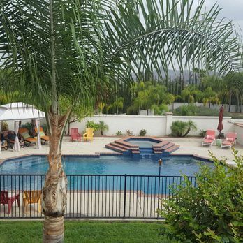 Backyard pool with a spa, surrounded by a black fence, palm trees, lounge chairs, and a gazebo.