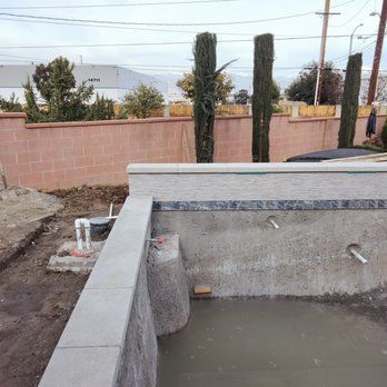 Partially constructed pool with concrete walls, near a brick wall and trees.