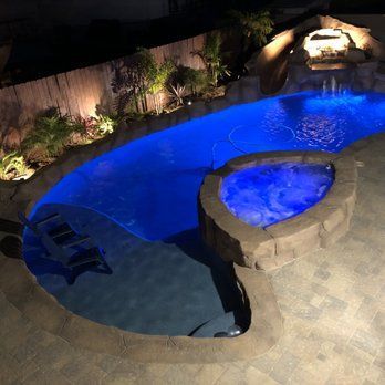 Night view of a blue lit pool and spa with waterfall feature surrounded by landscaping and rocks.