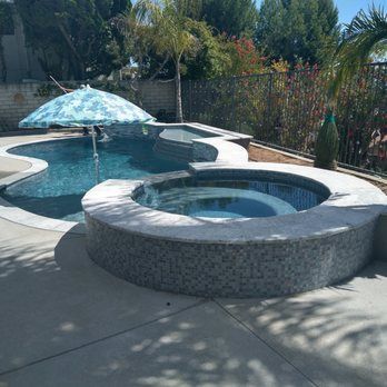A pool and jacuzzi with blue water, surrounded by stone and a concrete patio, umbrella overhead.