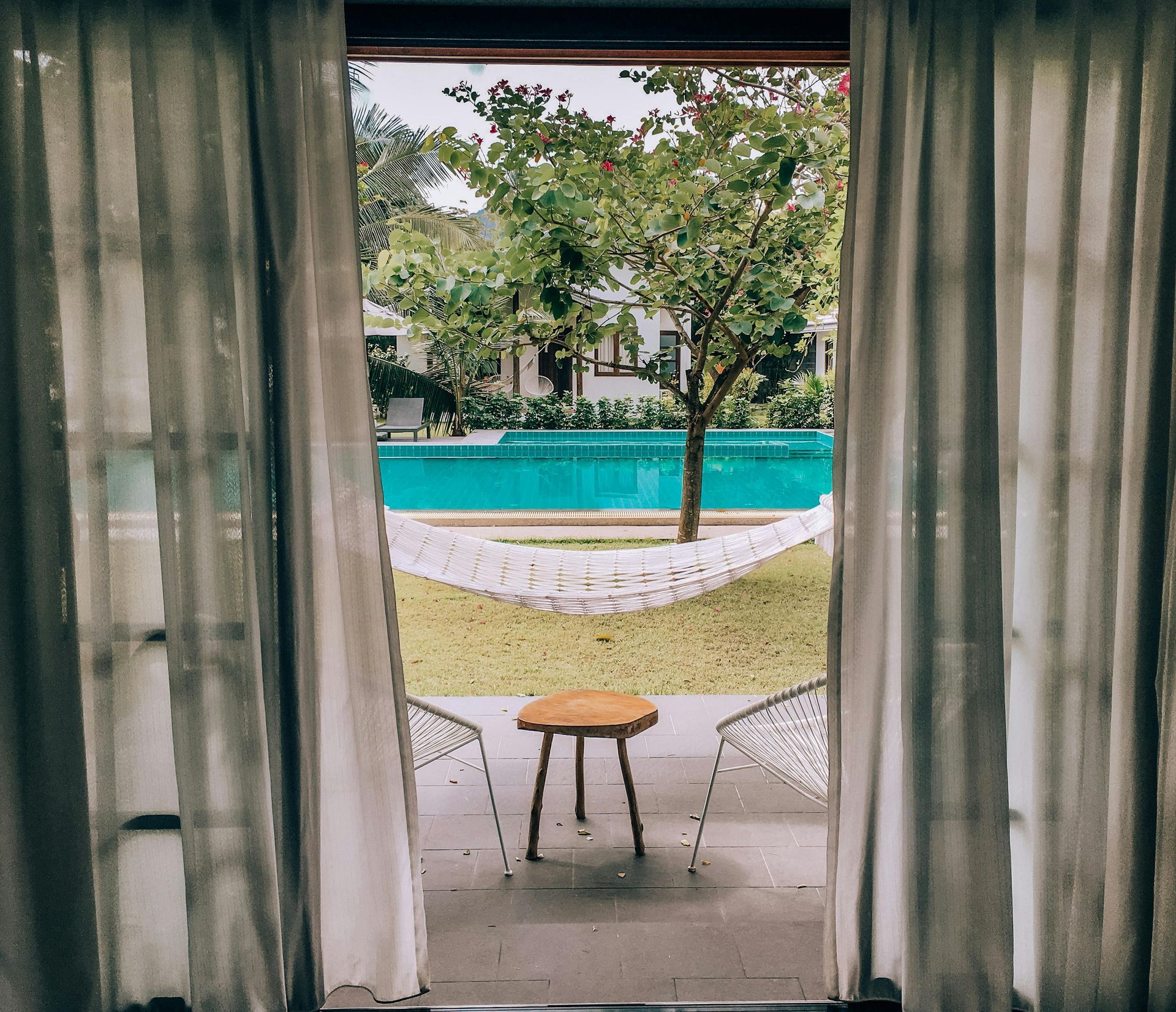 View of a pool and trees through open doorway framed by sheer curtains. A stool sits outside.