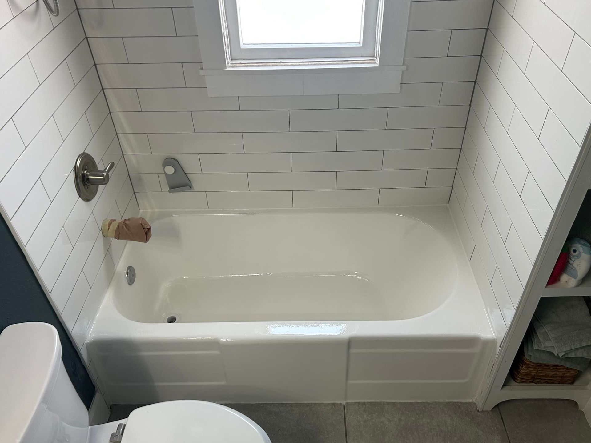 White bathroom with tub, toilet, and shelving. White subway tile on walls, small window above the tub.