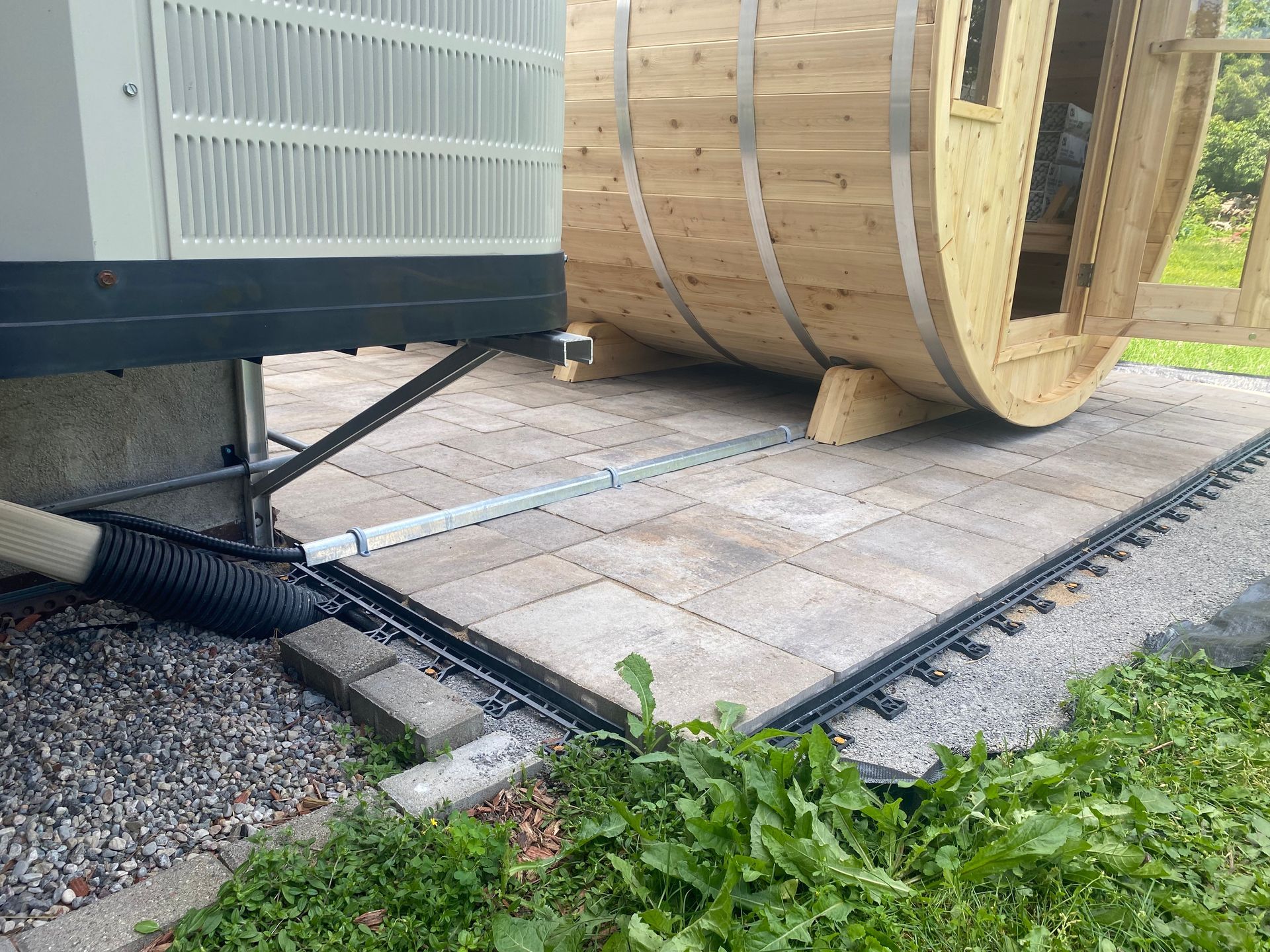 Outdoor scene: Barrel sauna next to an air conditioning unit on a paved patio with a toy train track.