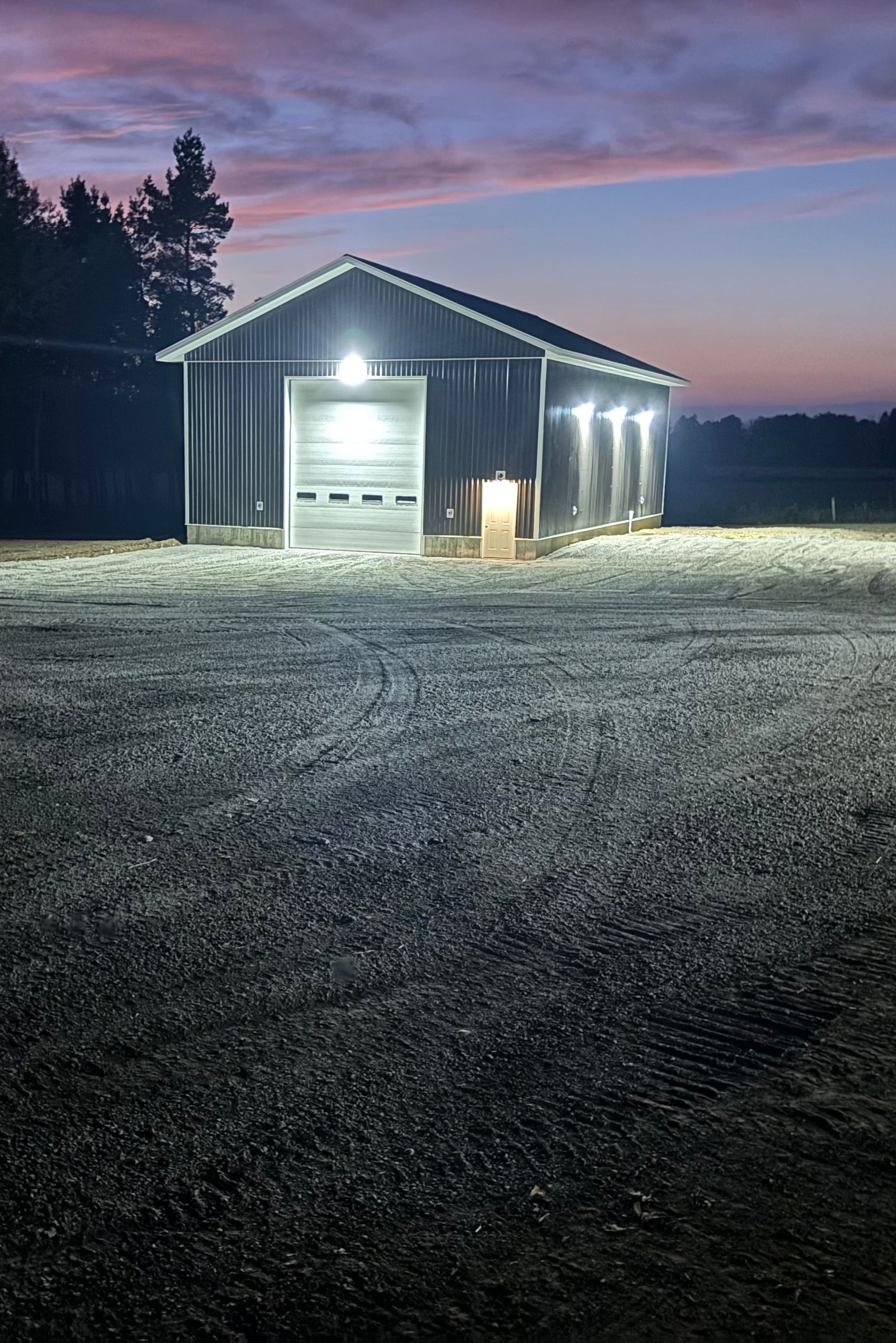 Barn illuminated at dusk, with gravel in the foreground and a colorful sky.