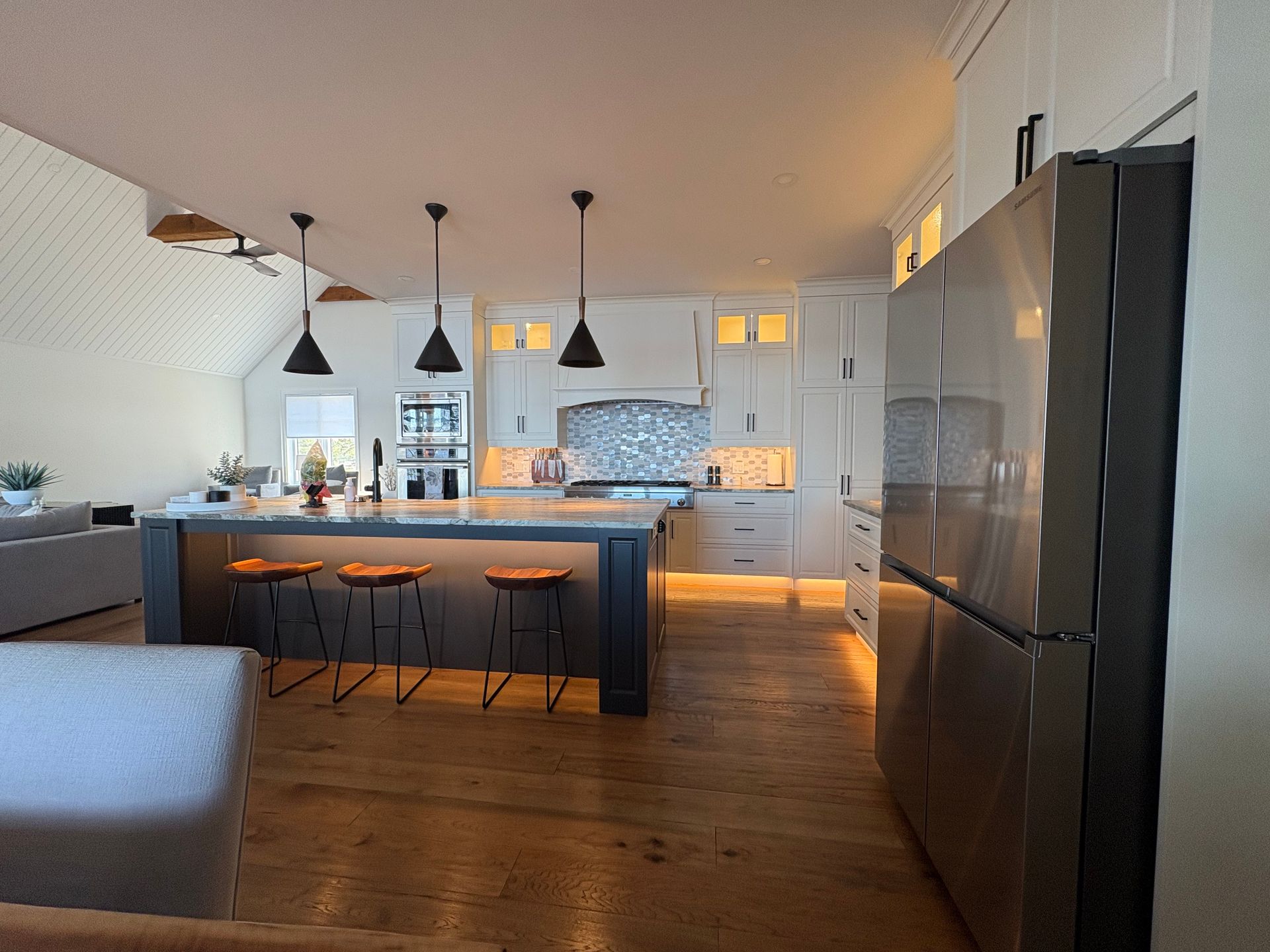 Modern kitchen with island, pendant lights, stainless steel refrigerator, and hardwood floors.