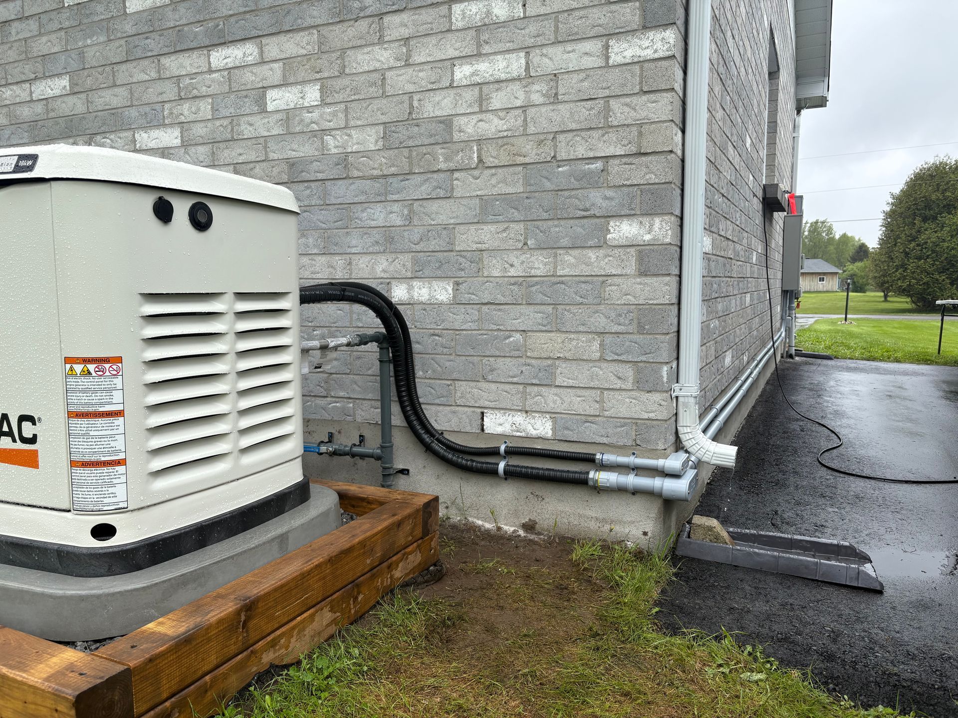 A standby generator next to a brick house. Black electrical conduits run along the wall.