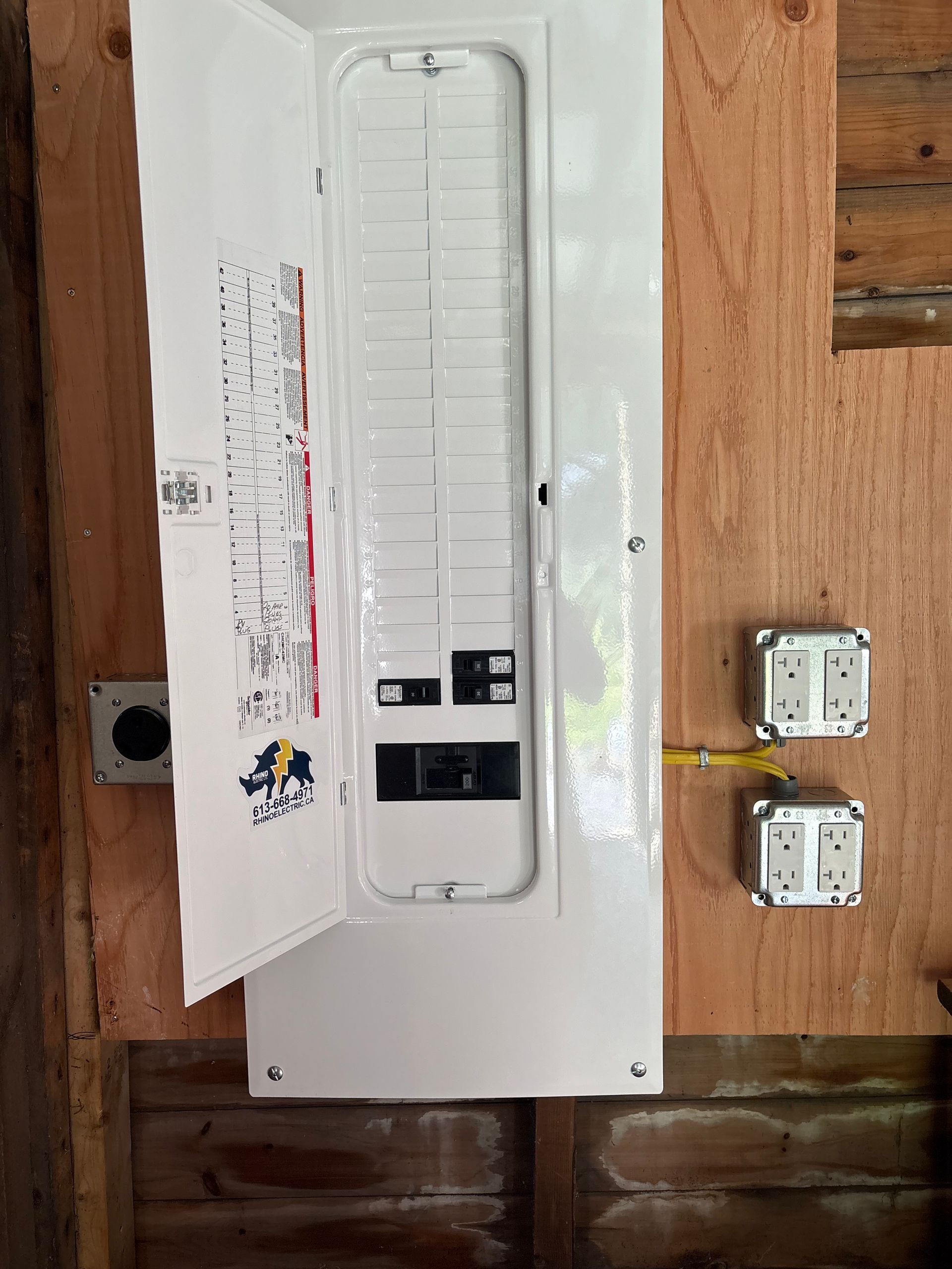 Opened electrical panel with circuit breakers, mounted on wood with two electrical outlets.