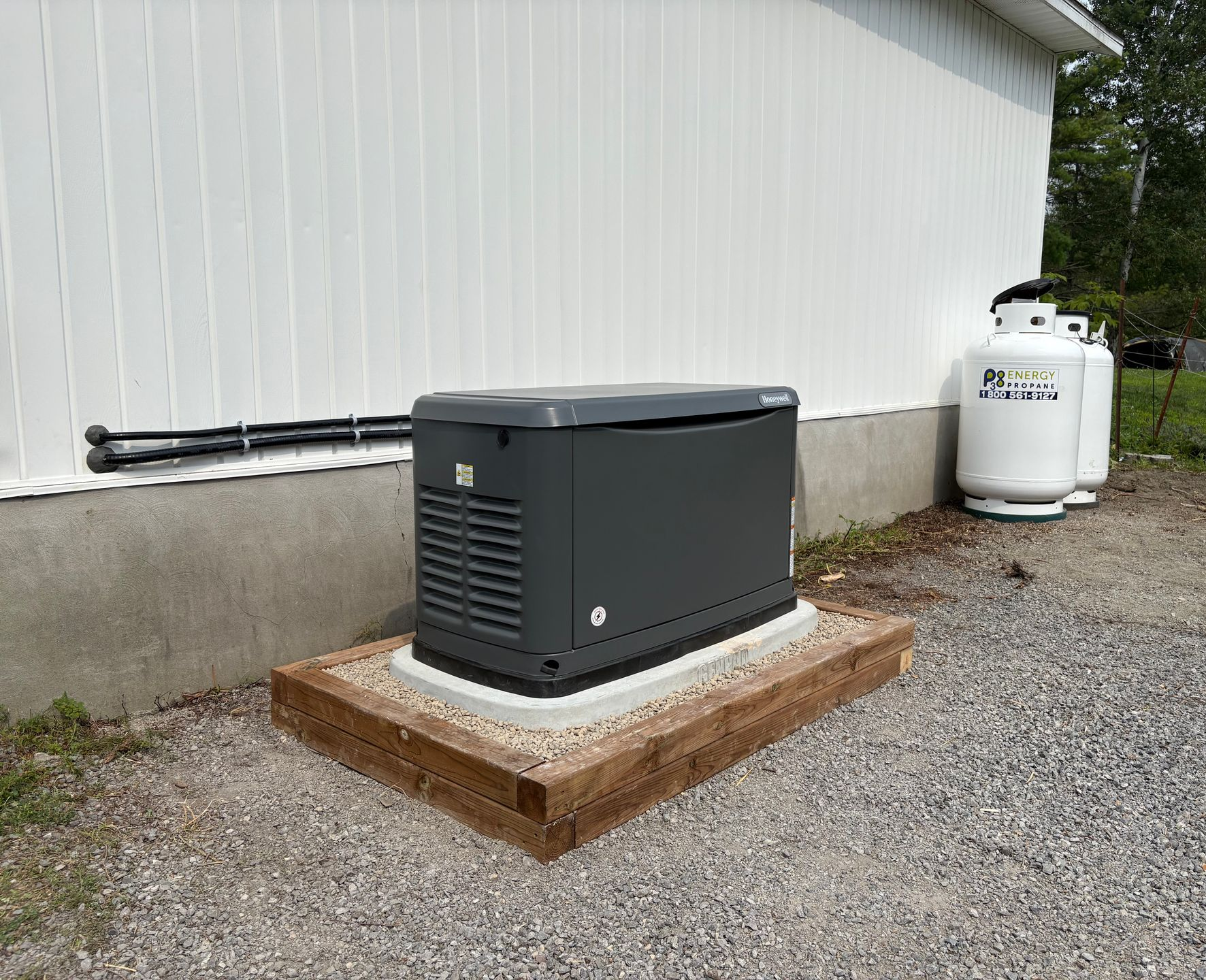 Gray generator on a concrete pad near a white building and propane tanks.