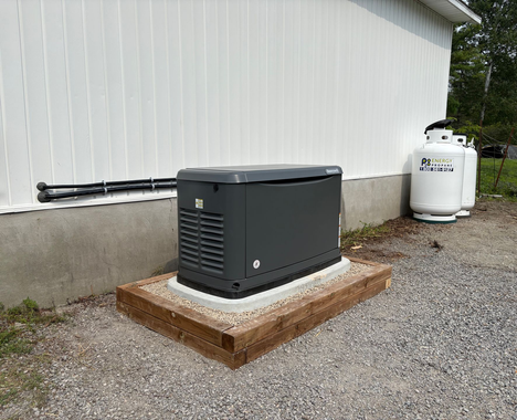 Gray generator on a concrete pad near a white building and propane tanks.