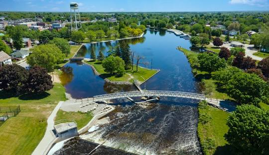 Aerial view of a river and dam in a town with lush green trees, a bridge, and a water tower.