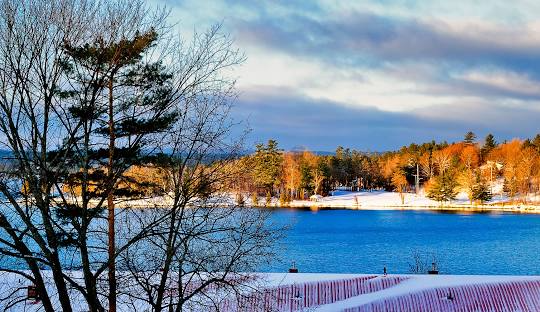 Snowy lake scene with trees, blue water, and fall foliage in the background.