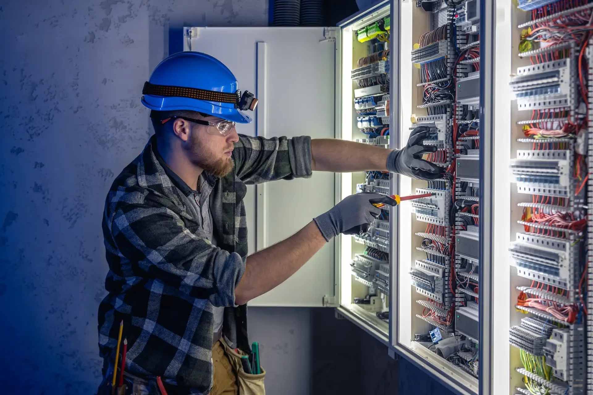 Electrician in blue hard hat, working on a control panel with tools.