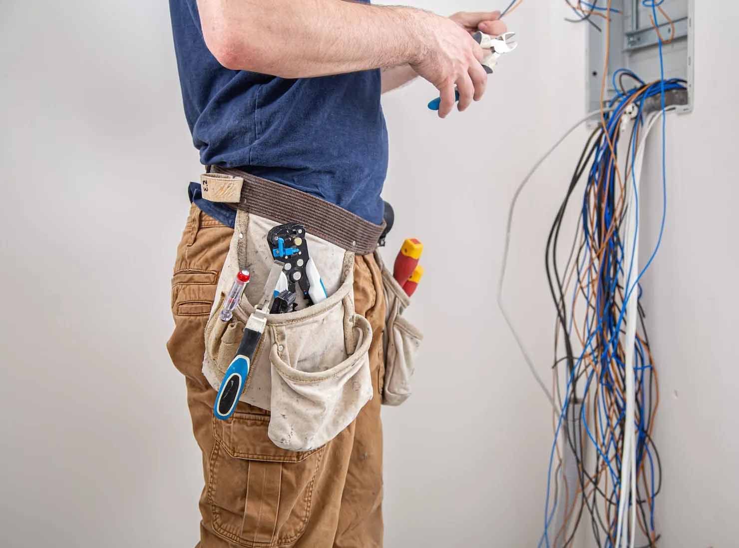 Electrician cutting wires near a wall-mounted panel. Wires are blue, white, and brown. He wears a tool belt and brown pants.