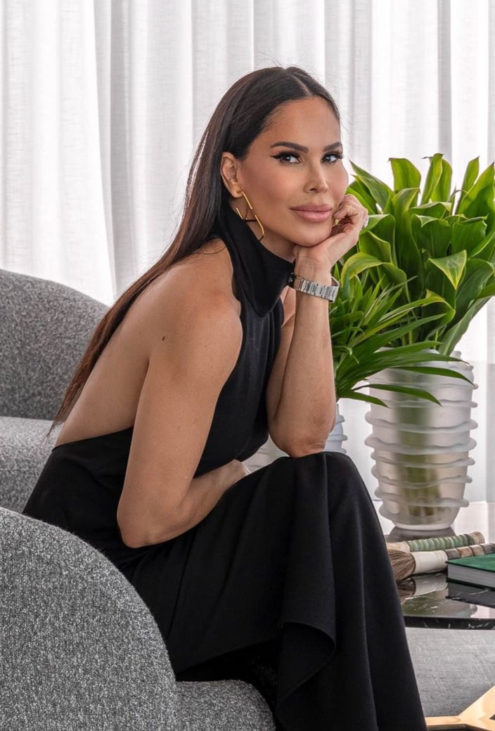 Uma mulher de vestido preto está sentada em uma cadeira ao lado de uma planta em vaso.