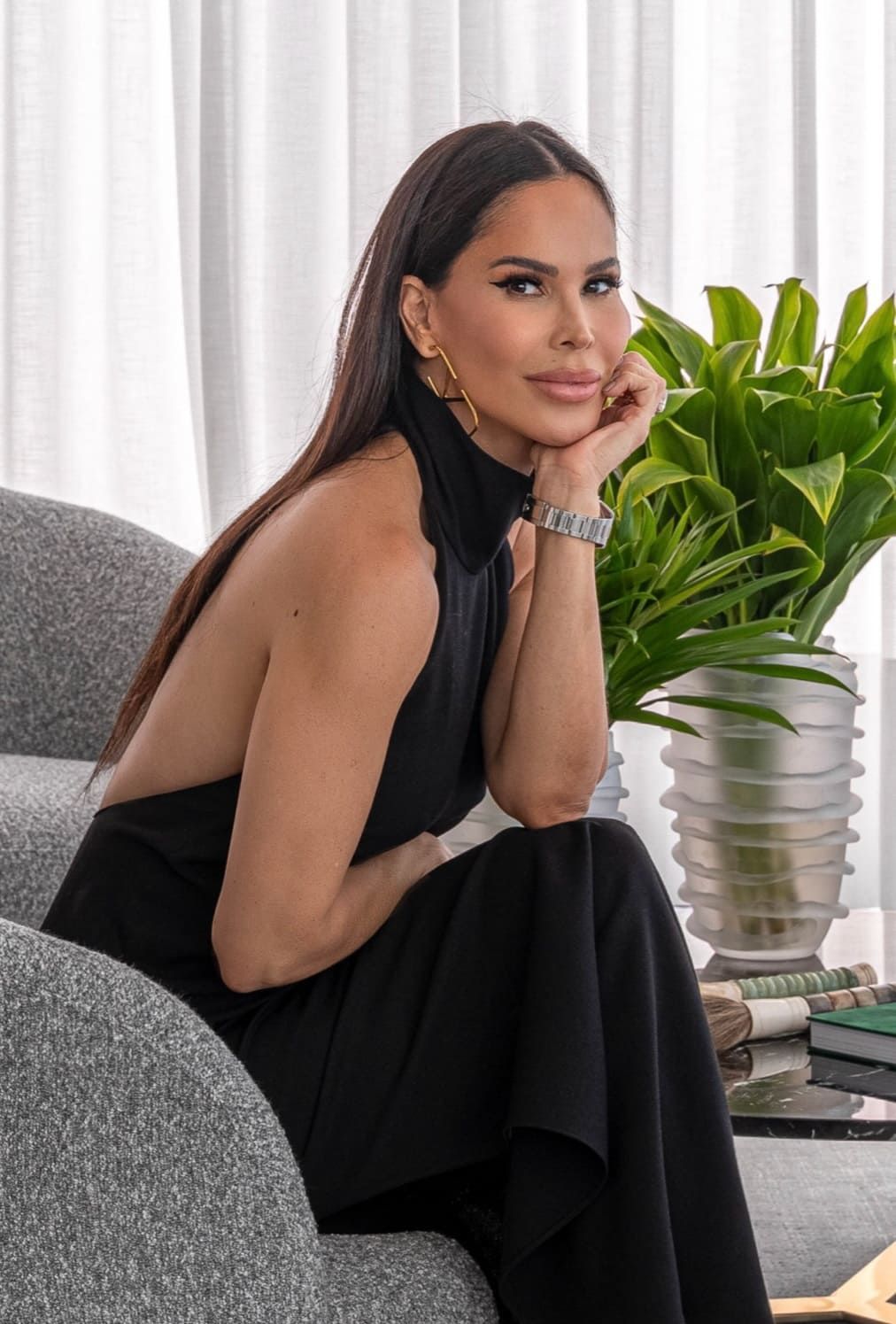 Uma mulher de vestido preto está sentada em uma cadeira ao lado de uma planta em vaso.