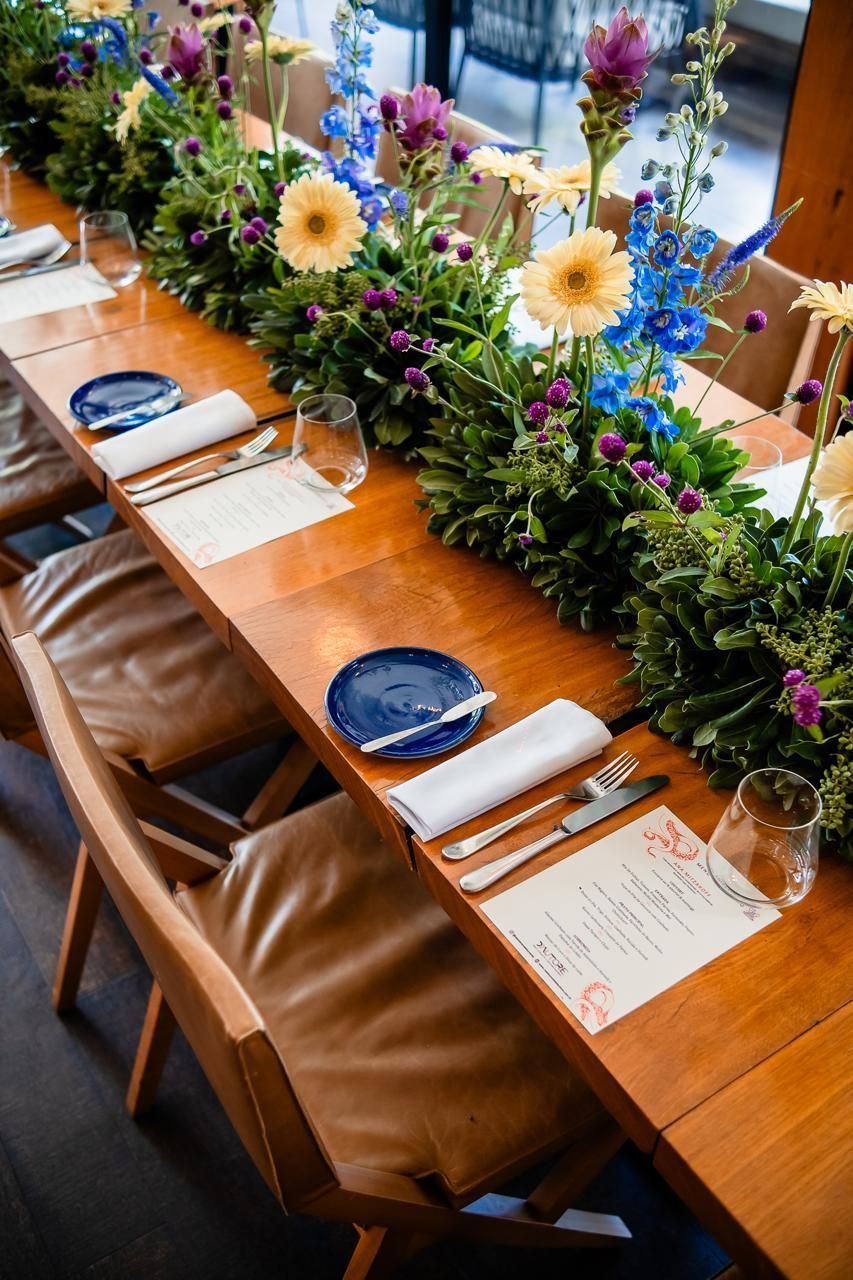 Uma longa mesa de madeira com pratos, talheres e flores sobre ela.