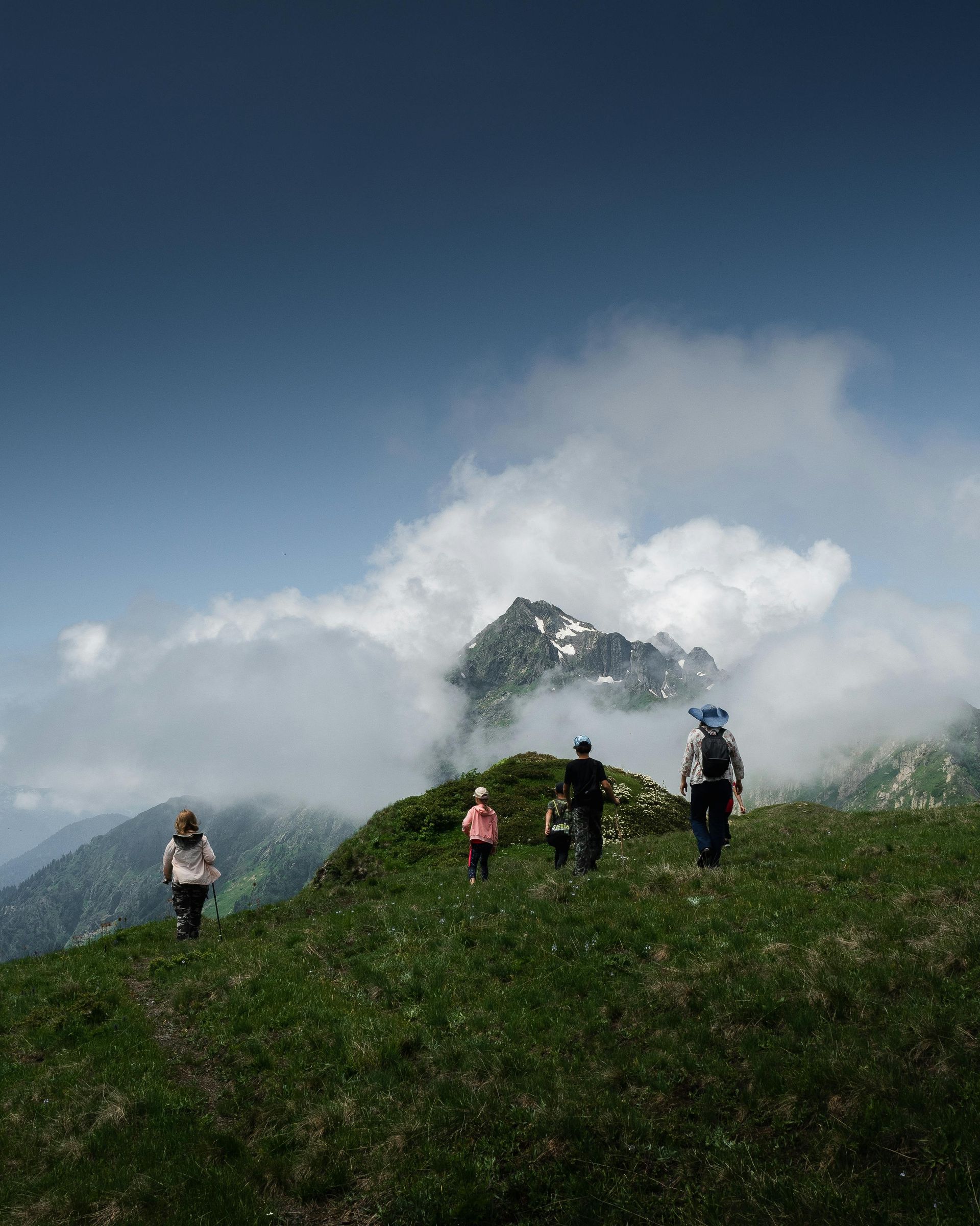 Hikers walking along a green mountain ridge above clouds under a blue sky