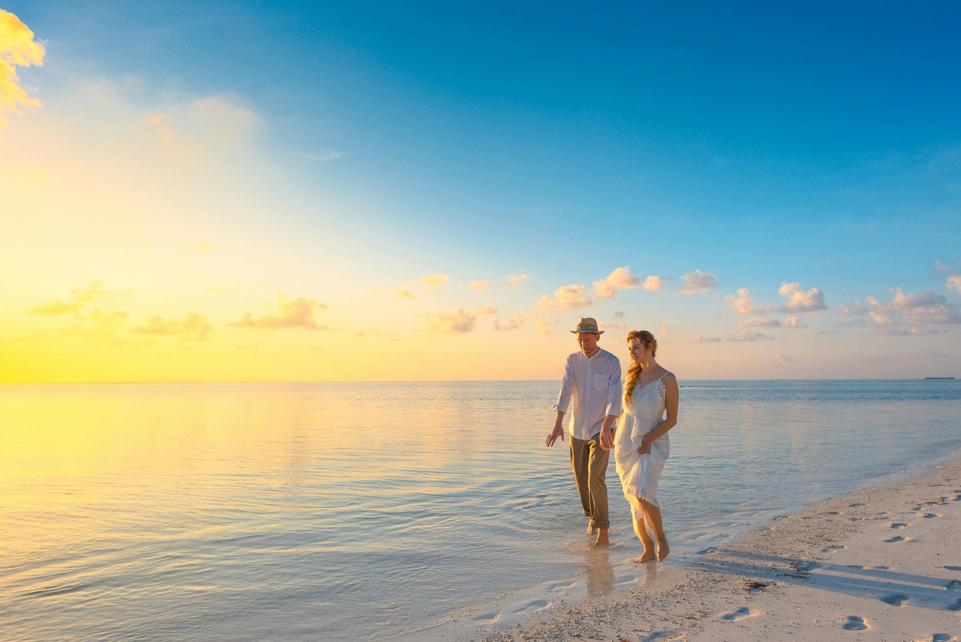 Una pareja camina de la mano por una playa tranquila e iluminada por el sol al amanecer, con la luz dorada reflejándose en el agua.